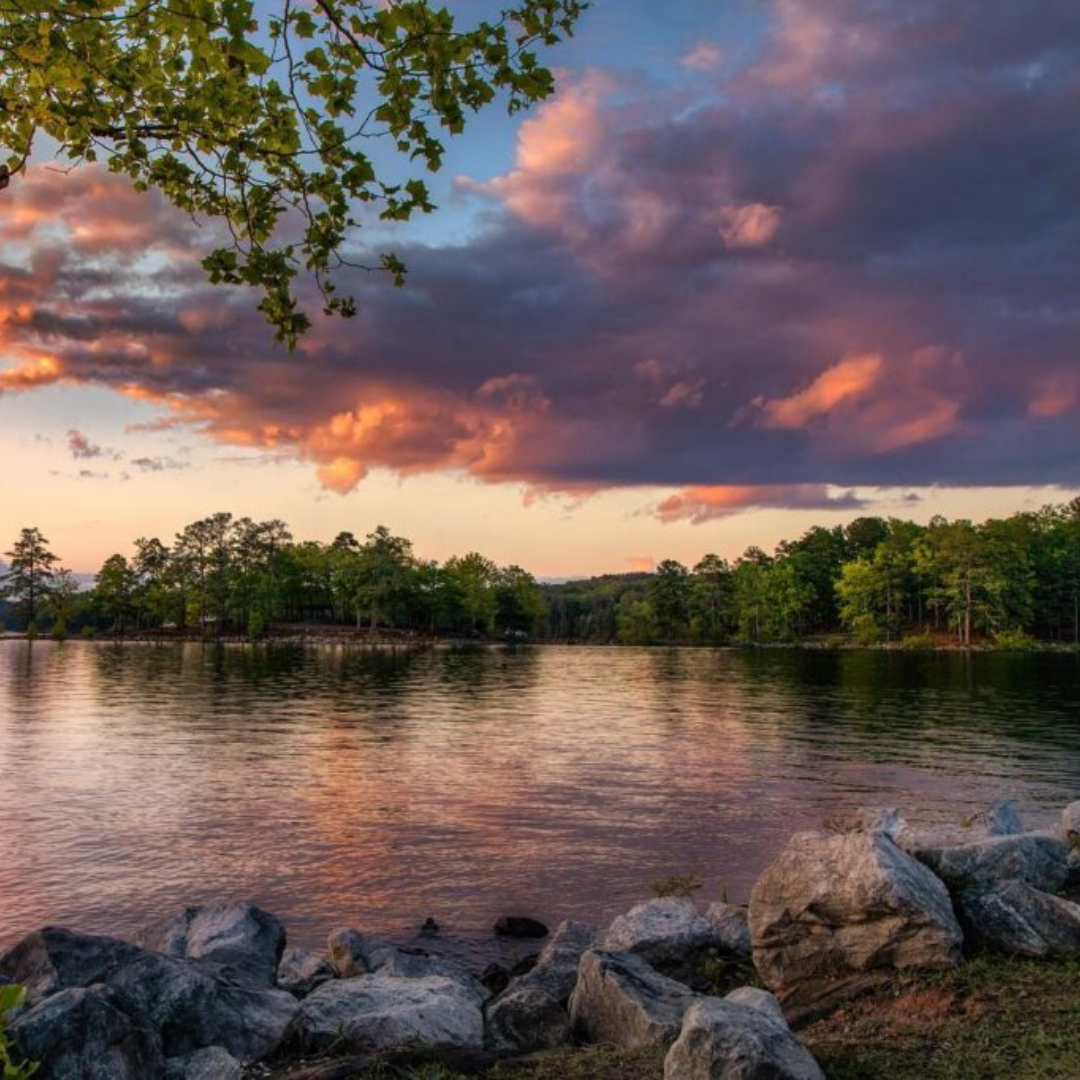 A scenic view of a lake at sunset in Acworth, with colorful clouds reflecting on the water, surrounded by trees and rocks in the foreground—a handful of reasons to put this enchanting spot on your travel bucket list.