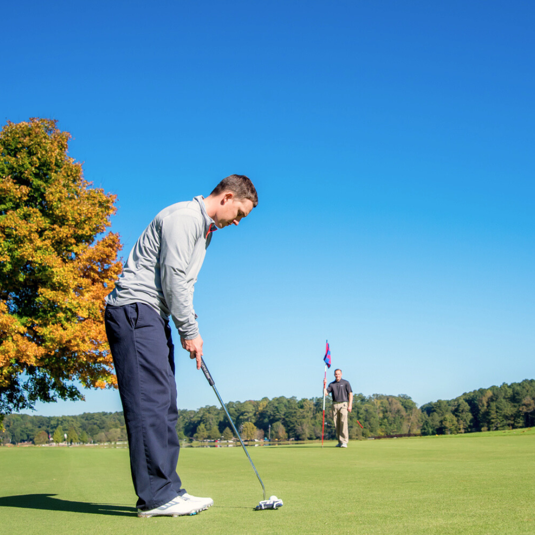 A golfer in mid-swing focuses on a putt on a green course, with Acworth's blue sky and fall-colored tree providing a picturesque backdrop. Another person stands near a flag, offering just one of the handful of reasons to put this charming town on your travel bucket list.