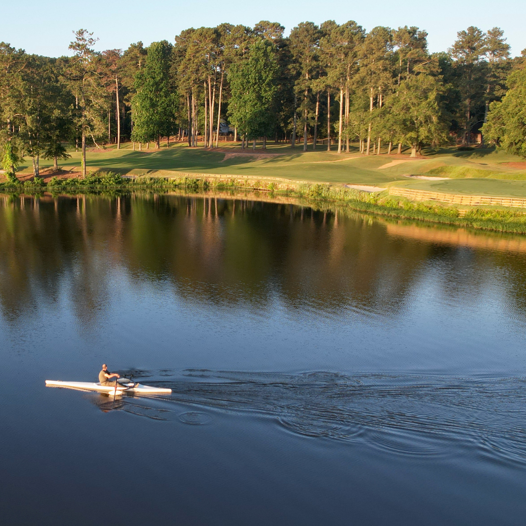 A person in a kayak paddles across a calm lake surrounded by trees and a grassy landscape, offering just one of a handful of reasons to put Acworth on your travel bucket list.