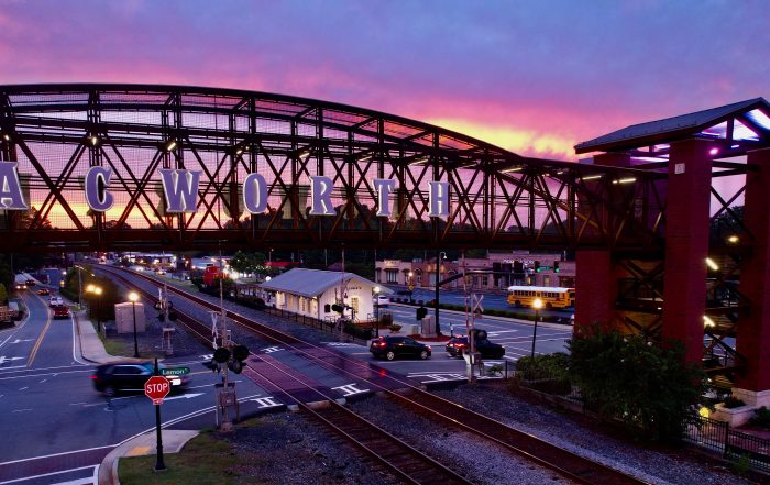 Pedestrian bridge labeled "ACWORTH" spans over railroad tracks and a road at sunset, offering a glimpse of cars and a stop sign below—just one of A Whole Handful of Reasons to put Acworth on your Travel Bucket List.