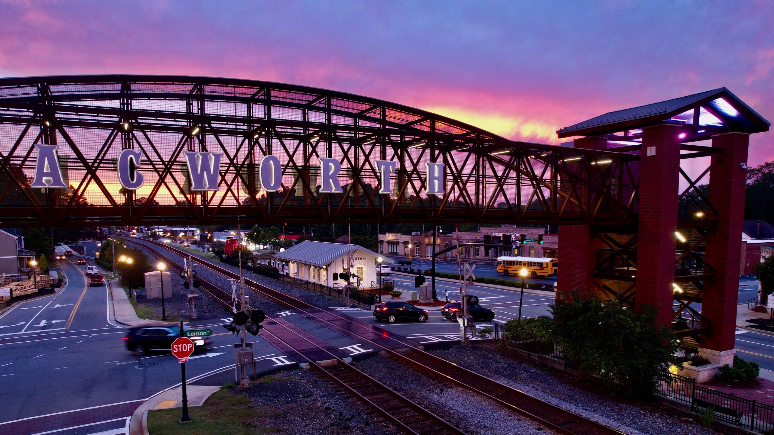 Pedestrian bridge labeled "ACWORTH" spans over railroad tracks and a road at sunset, offering a glimpse of cars and a stop sign below—just one of A Whole Handful of Reasons to put Acworth on your Travel Bucket List.
