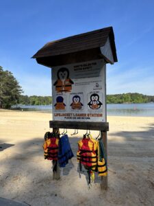 A lakeside lifejacket loaner station with several lifejackets hanging below a sign displaying water safety rules and instructions.
