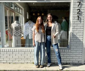 Two women stand smiling outside a storefront with a sign reading "Women's Club" in Downtown Acworth. Mannequins wearing dresses are visible in the window behind them, capturing the vibrant charm of Cafe de Coco.