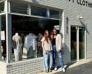 Two women stand smiling outside a white brick storefront in Downtown Acworth, with a large window displaying mannequins and a sign reading "WOMENS CLOTHING," just steps away from the great wine market in town.