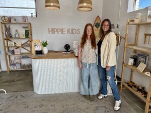 Two women stand smiling behind the counter of a shop called "Hippie Kids," surrounded by shelves with various items and decor in a bright, modern space, just steps from the great wine market in Downtown Acworth.