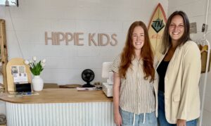 Two women stand smiling behind the front counter of Hippie Kids Cafe de Coco, with a vase of white tulips and a surfboard in the background at their cozy Downtown Acworth shop.