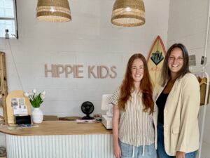Two women stand smiling behind a wooden counter in the Hippie Kids boutique with surfboard decor and a "HIPPIE KIDS" sign on the wall, capturing the relaxed vibe of Downtown Acworth.