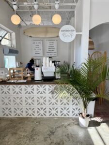 A small coffee shop counter with a patterned front, espresso machine, baked goods display, and a potted palm; menu and sign read "Café de Coco," welcoming hippie kids in downtown Acworth.