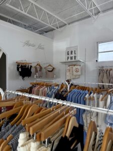 Clothing racks with assorted garments in a boutique store; a shelf with bags, a "GRL PWR" sign, and a window evoke the vibrant spirit of Hippie Kids Cafe de Coco in Downtown Acworth.