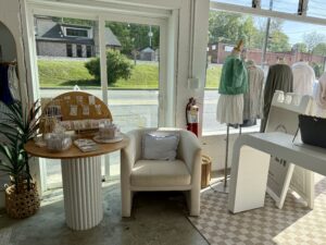 A small boutique shop interior in Downtown Acworth with a display table of jewelry, a white armchair, a fire extinguisher, and clothing on mannequins near large windows—perfect for exploring after visiting Hippie Kids Cafe de Coco.