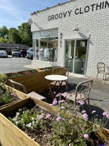 Outdoor seating area with a round table and metal chairs surrounded by flower planters in front of "Groovy Clothing," a white brick store near Downtown Acworth—perfect for relaxing after shopping at this hippie-inspired spot.