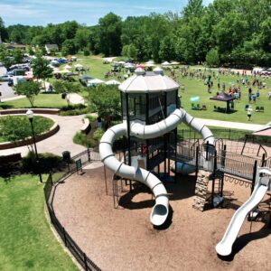 A large playground with spiral slides is in the foreground, while people gather under tents and on the grass in a park setting in the background during the 2025 Pro Wakeboard Tour.