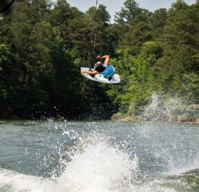 A person wearing a blue shirt performs an aerial trick on a wakeboard over a lake during a weekend in Acworth, with trees and water spray in the background.