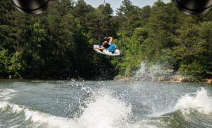 A person wearing a blue shirt performs an aerial trick on a wakeboard over a lake during a weekend in Acworth, with trees and water spray in the background.