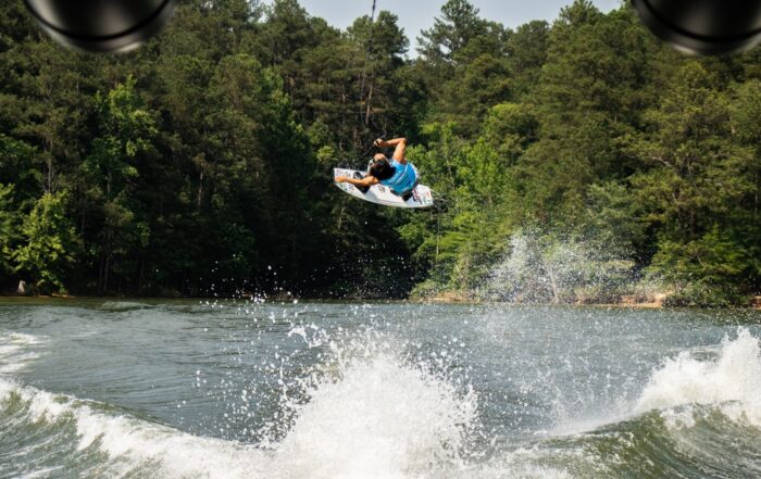 A person wearing a blue shirt performs an aerial trick on a wakeboard over a lake during a weekend in Acworth, with trees and water spray in the background.