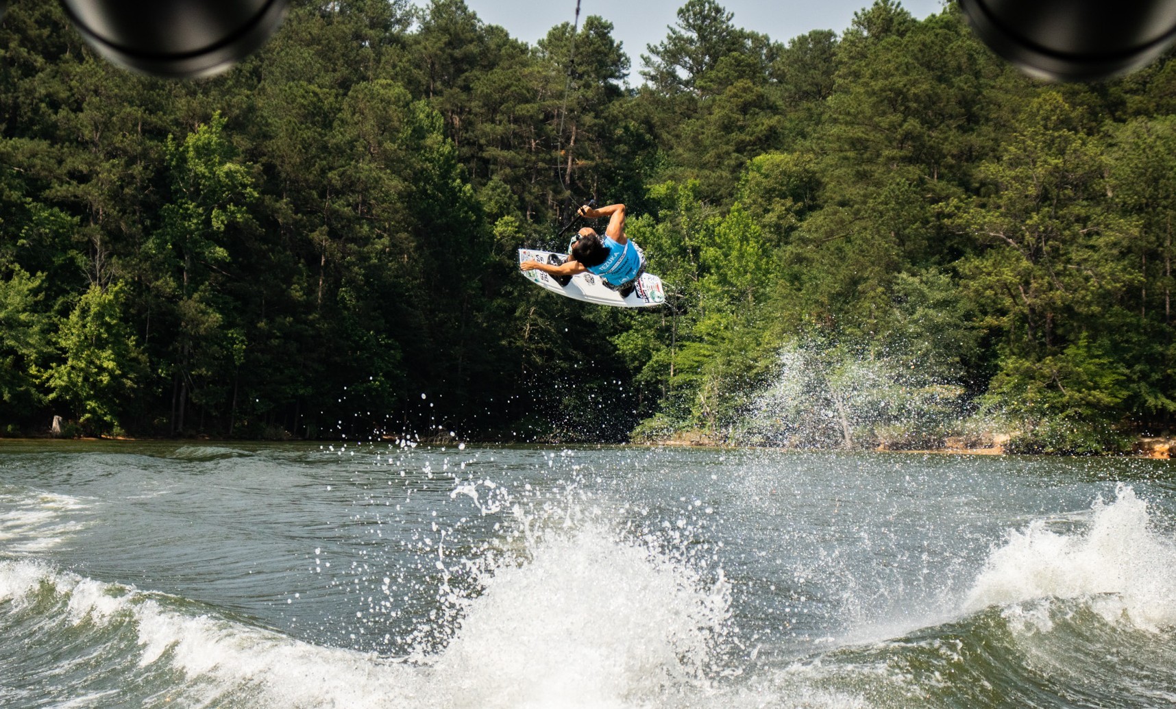 A person wearing a blue shirt performs an aerial trick on a wakeboard over a lake during a weekend in Acworth, with trees and water spray in the background. A person wearing a blue shirt performs an aerial trick on a wakeboard over a lake during a weekend in Acworth, with trees and water spray in the background.