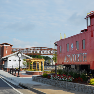 A bright red train caboose labeled "Acworth" is displayed near a flower bed, with a pedestrian bridge and a small white building in the background—capturing the charm of Acworth, home to events like the 2025 Pro Wakeboard Tour.