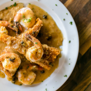 A white plate with fried breaded fish topped with shrimp and a creamy sauce, garnished with chopped herbs, makes the perfect dish for a weekend in Acworth, served on a rustic wooden table.