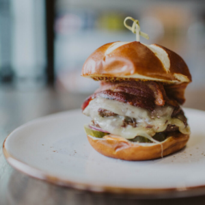 A cheeseburger with melted cheese, bacon, pickles, and a pretzel bun is served on a white plate—the perfect bite for your weekend in Acworth.