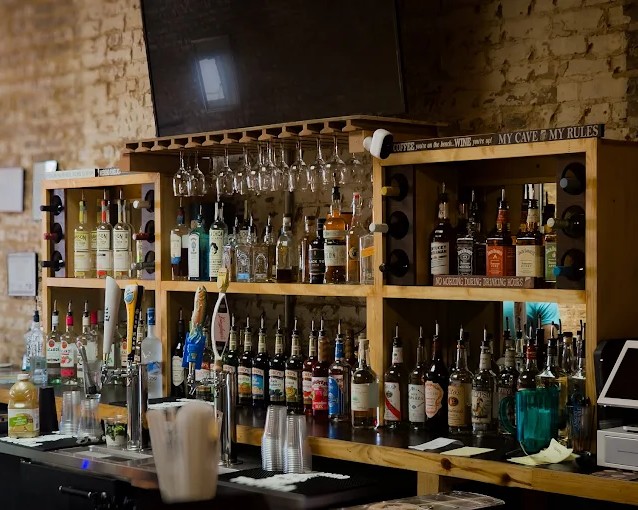 A bar with a variety of liquor bottles, wine glasses hanging above, plastic cups, and a cash register on a wooden counter against a brick wall.