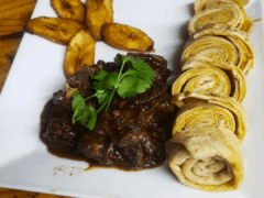 A square white plate with fried plantain slices, rolled flatbread, and a serving of stewed meat garnished with cilantro.