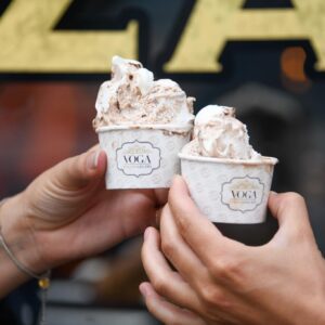 Two hands hold up cups of swirled gelato labeled "Yoga Italian Gelato," showcasing one of the tastiest Ways to Cool Off this Summer in Acworth against a softly blurred background.
