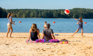 Two adults relax on beach towels, watching three children play with a beach ball on a sandy lakeshore—just one of A Whole Handful of Reasons to put Acworth on your Travel Bucket List.