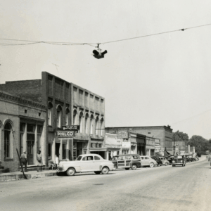 Black-and-white photo of a small town street with parked vintage cars, brick storefronts, and a traffic signal suspended overhead. Learn more about historic Acworth locations captured in timeless moments like this.