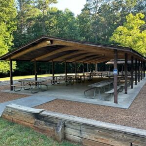 A covered outdoor pavilion with multiple picnic tables on a concrete slab offers one of the best Ways to Stay Cool this Summer in Acworth, surrounded by trees and a low wooden barrier for added shade and comfort.