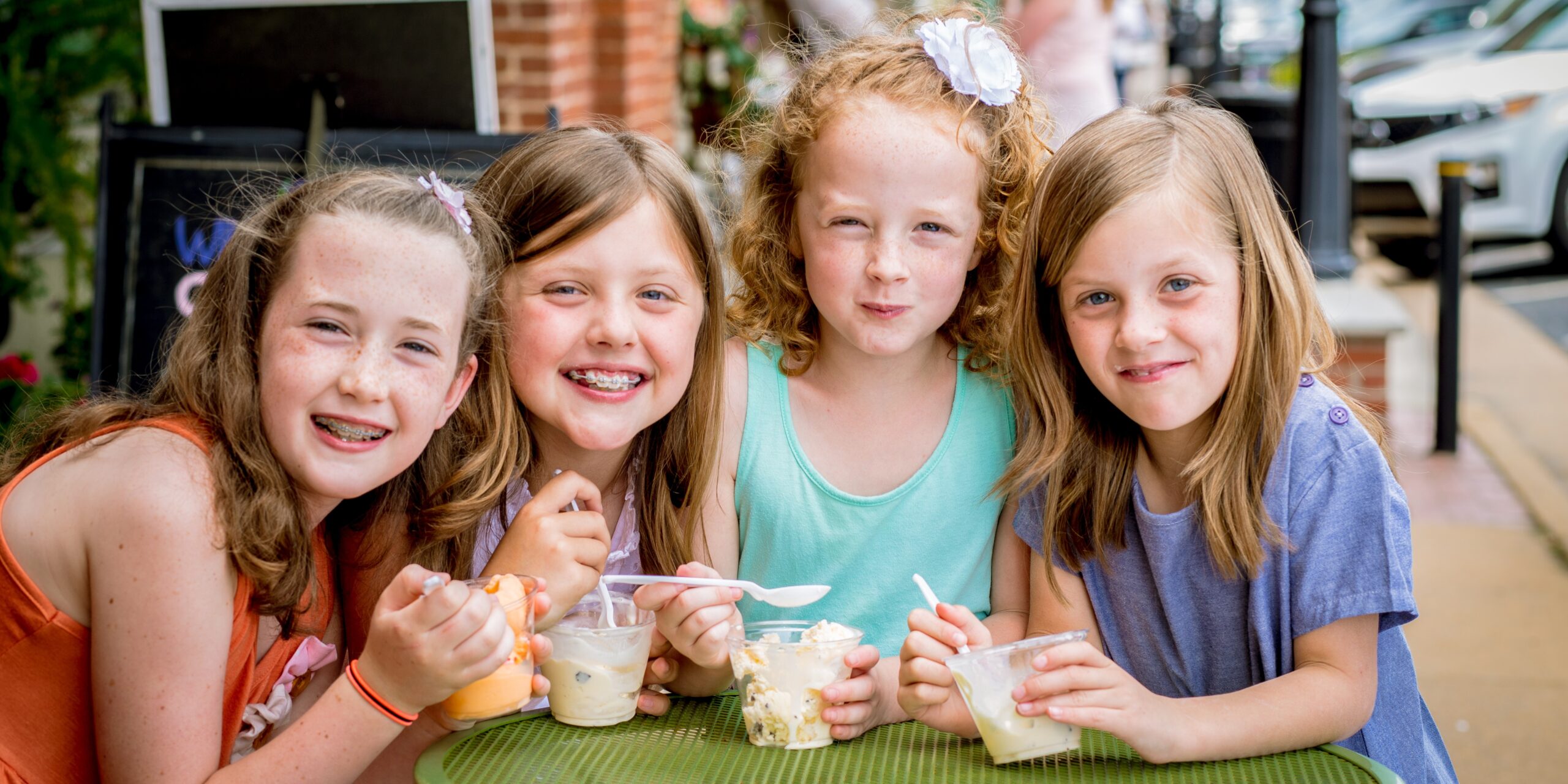 Four young girls sit at an outdoor table, smiling and eating ice cream from cups with spoons on a sunny day—one of the best Ways to Stay Cool this Summer in Acworth. Four young girls sit at an outdoor table, smiling and eating ice cream from cups with spoons on a sunny day—one of the best Ways to Stay Cool this Summer in Acworth.