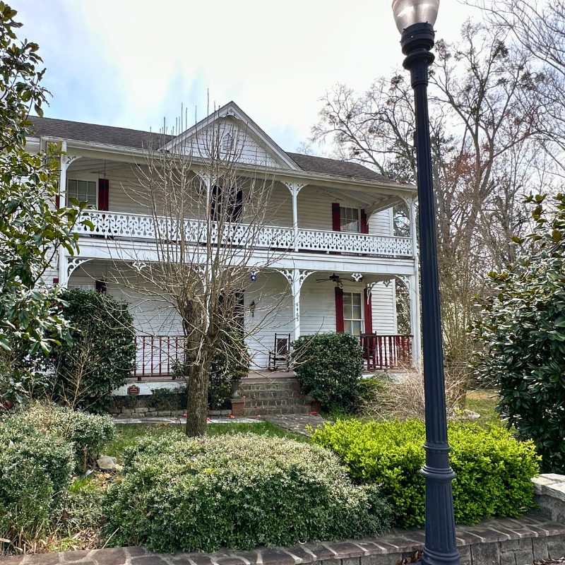 A two-story white house with a covered porch and red railings, surrounded by bushes and trees, with a streetlamp in the foreground. Learn more about historic Acworth locations like this charming residence. A two-story white house with a covered porch and red railings, surrounded by bushes and trees, with a streetlamp in the foreground. Learn more about historic Acworth locations like this charming residence.