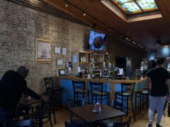 A modern bar with wooden tables and chairs, a brick wall, a TV displaying food, and a stained glass skylight overhead. Two men are in the room, one near a table and one walking toward the bar.