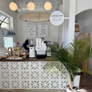 A modern coffee bar with a patterned counter, coffee machine, stacked cups, menu board, and a barista working—one of the best Ways to Stay Cool this Summer in Acworth. A potted palm plant adds a refreshing touch in the foreground.