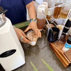A person places a lid on a cup of iced coffee at a counter with various syrups, a grinder, and coffee-making tools—one of the tastiest ways to stay cool this summer in Acworth.