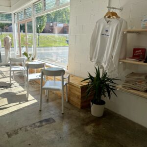 Bright cafe corner with white chairs, wooden tables, potted plant, and a sweatshirt on the wall. Large windows let in natural light—one of the cozy ways to stay cool this summer in Acworth while enjoying a relaxing afternoon indoors.