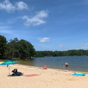 People relax on a sandy beach beside a lake in Acworth with an umbrella, towels, and inflatables; several people swim under a blue sky with scattered clouds—one of the best ways to stay cool this summer in Acworth.