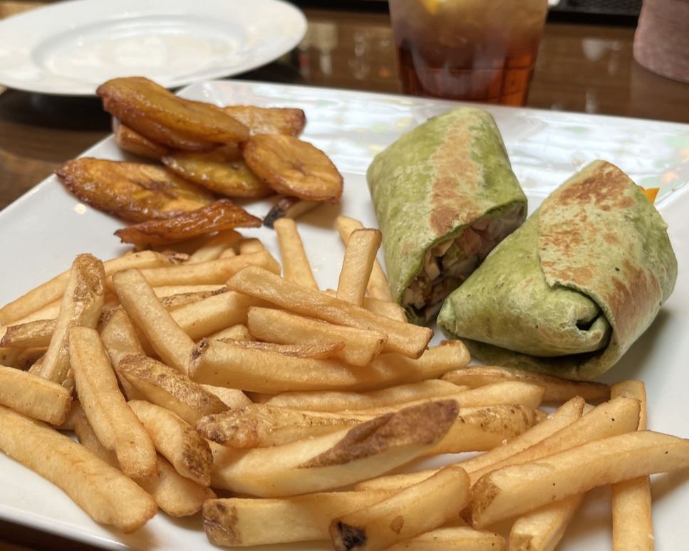 A plate with French fries, fried plantains, and a spinach tortilla wrap cut in half, next to a drink and an empty plate in the background.