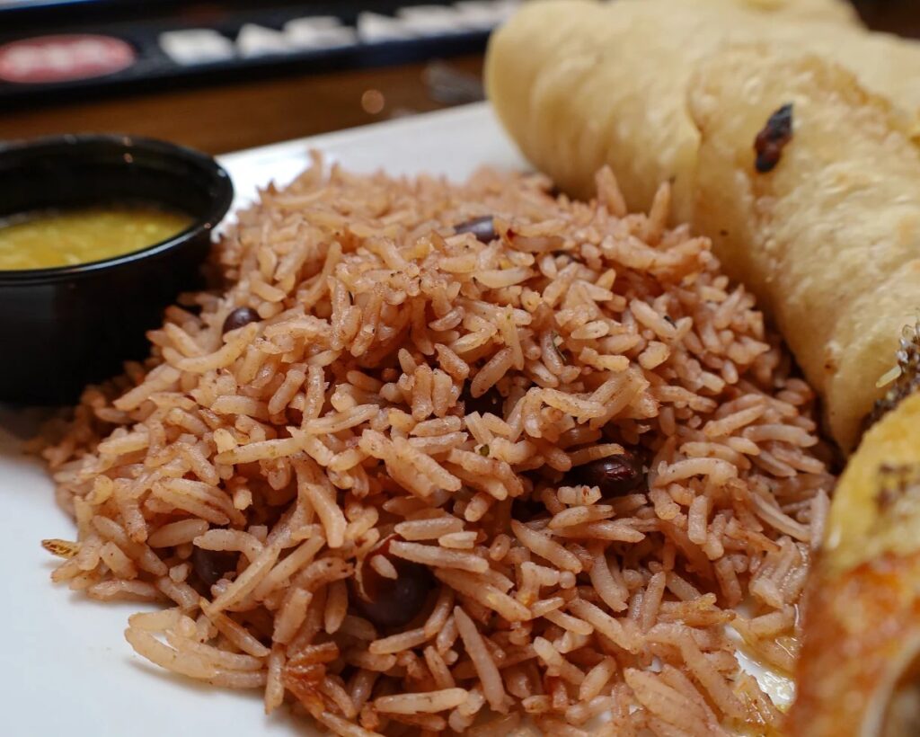 A plate with a serving of rice and beans, a piece of flatbread, and a small container of yellow sauce.