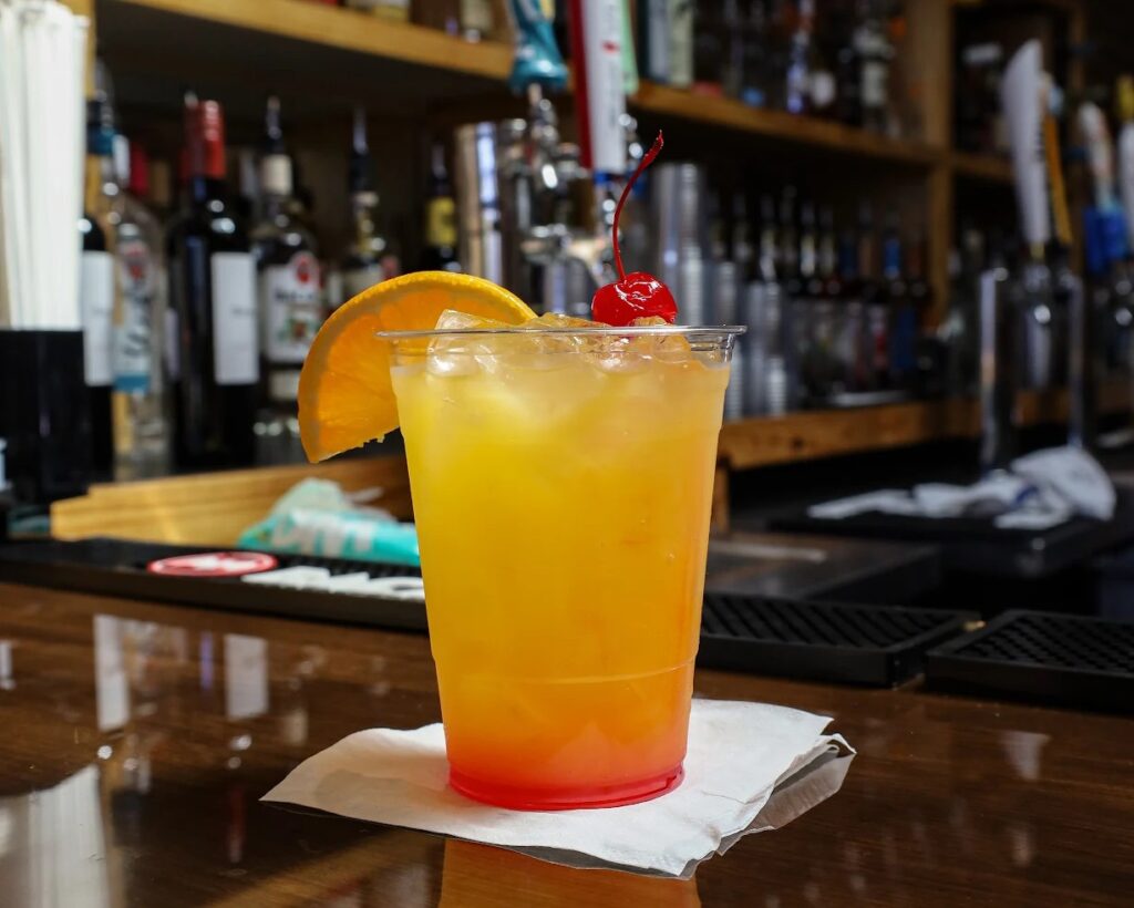 A plastic cup filled with an orange-yellow cocktail, garnished with an orange slice and a cherry, sits on a napkin on a bar counter with bottles in the background.