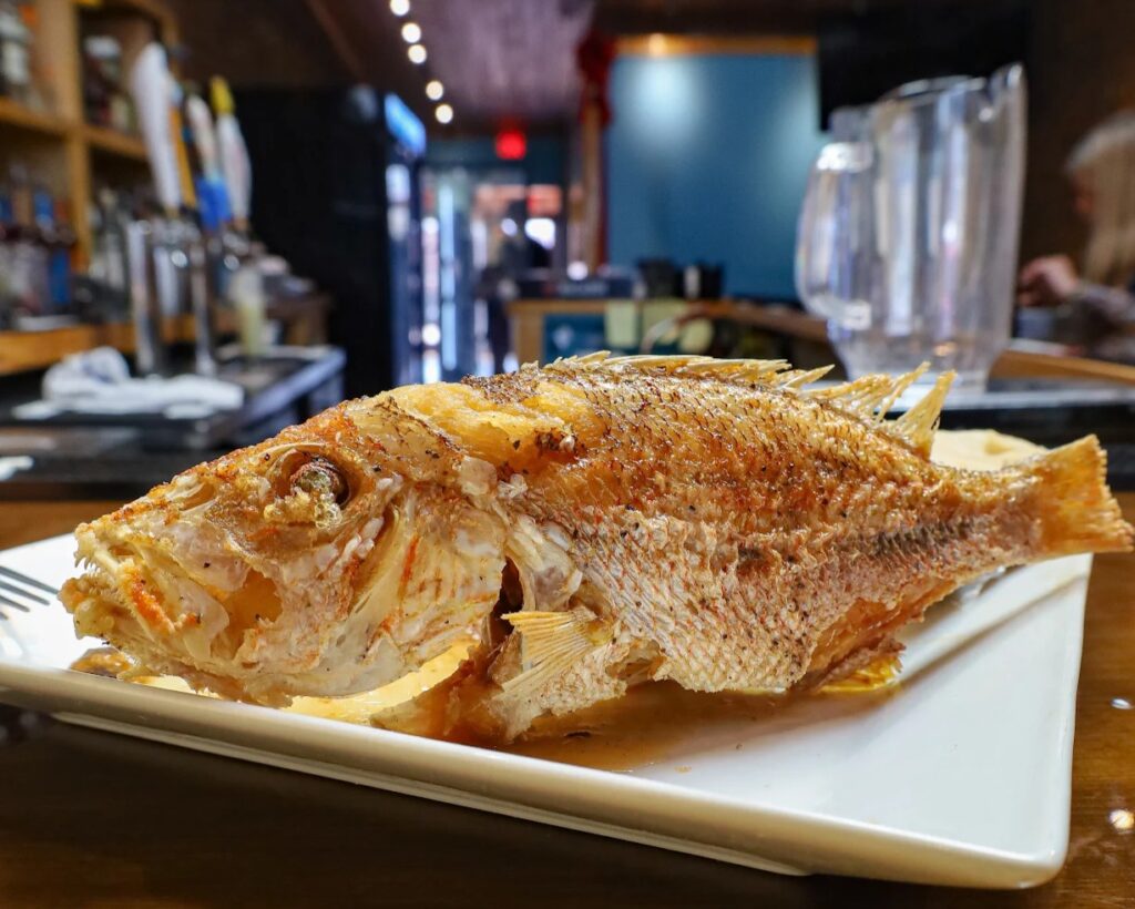 A whole cooked fish, seasoned and served on a white rectangular plate, sits on a wooden table in a restaurant setting.