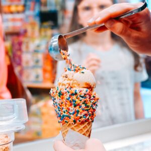 A hand holds an ice cream cone covered in rainbow sprinkles as another hand adds more sprinkles with a scoop—just one of the fun Ways to Stay Cool this Summer in Acworth. In the background, a person watches the treat take shape.