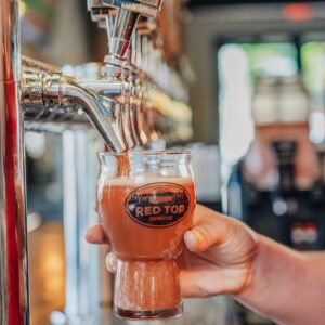 A hand holds a glass with the "Red Top Brewhouse" logo, filling it with beer from a tap at a bar—stay cool in Acworth with a refreshing local brew.