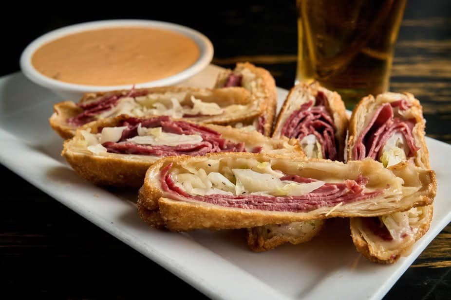 Sliced Reuben egg rolls filled with corned beef and sauerkraut on a white plate, served with a side of creamy dipping sauce and a glass of beer.