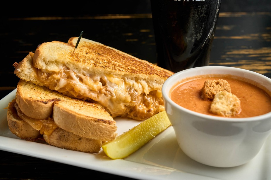 A grilled cheese sandwich is served with a bowl of tomato soup topped with croutons, a pickle spear, and a dark beverage in the background.