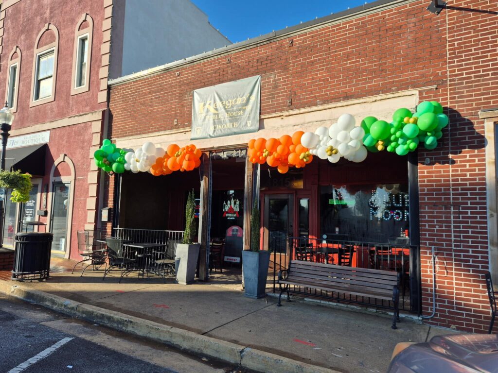 A brick storefront decorated with orange, white, and green balloons above the entrance, with empty patio seating and a sign above the door.
