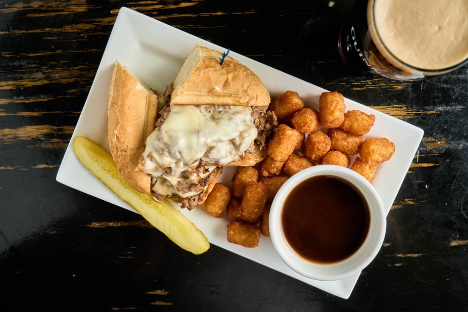 A sandwich with melted cheese, tater tots, a pickle spear, and a cup of brown dipping sauce are served on a rectangular white plate with a glass of dark beverage nearby.