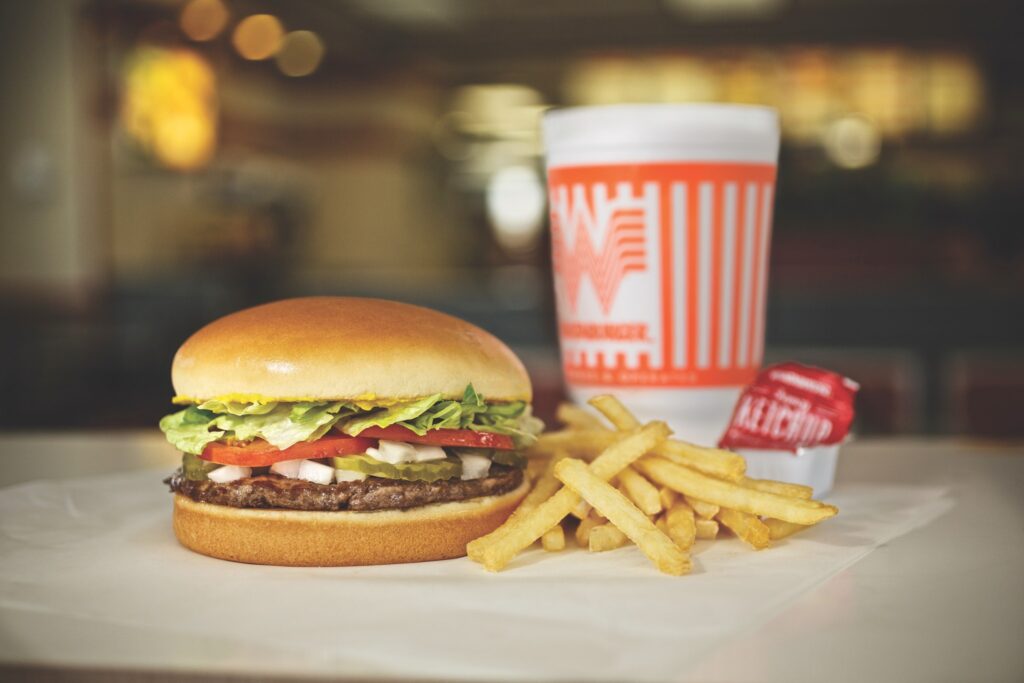 A hamburger with lettuce, tomato, and cheese is served with French fries, a condiment packet, and a branded soft drink cup on a table in a fast-food restaurant.