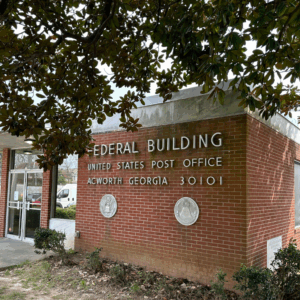 Red brick building with "Federal Building United States Post Office Acworth Georgia 30101" on the wall, two official seal plaques, and a glass entrance door visible on the left. Learn more about historic Acworth locations at this iconic site.