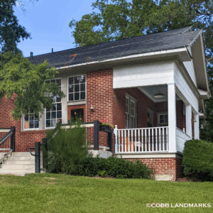 A small red brick house with white trim, front porch, and surrounding greenery under a clear blue sky—discover its charm as you Learn More About Historic Acworth Locations.