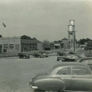 Black and white photo of vintage cars parked in a lot near industrial buildings and a tall water tower; an American flag is visible on the left. Learn more about historic Acworth locations captured in timeless moments like this.